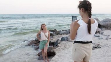 Tırpanlı ve cep telefonlu bir kız deniz kıyısındaki bir taşın üzerinde oturan annesinin fotoğraflarını çekiyor.