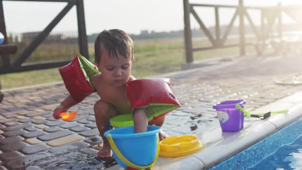 Boy with armbands playing with toys near the pool with clear water on the background of a summer sunset — Stock video