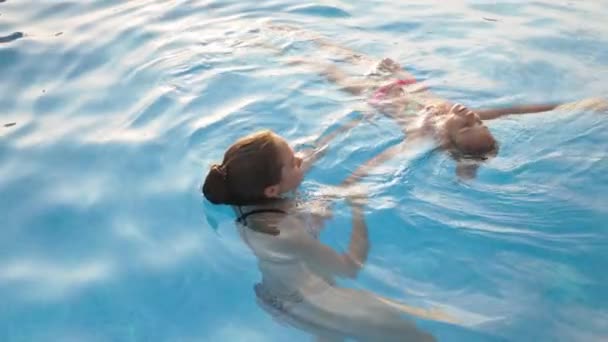 Bir abla küçük kız kardeşine pembe mayo içinde temiz su havuzunda yüzmeyi öğrenmesi için yardım eder. — Stok video