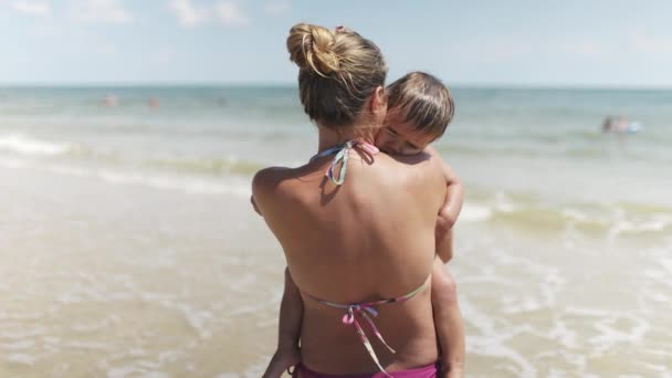 Mama w letnim bikini trzyma dziecko, które chce spać na plaży nad morzem na wakacjach — Wideo stockowe