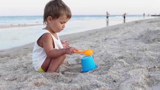 Jongen spelen met speelgoed op het strand gebouw kralen en torentjes glimlachen naar iemand achter de schermen op zomervakantie — Stockvideo