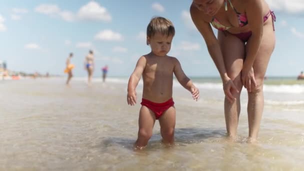 Ребенок собирает раковины и гальку в море на песчаном дне со своей матерью под летним солнцем в отпуске — стоковое видео
