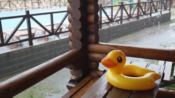 Um pato inflável encontra-se na mesa de madeira no gazebo no centro de recreação contra o pano de fundo da piscina coberta com chuva — Vídeo de Stock
