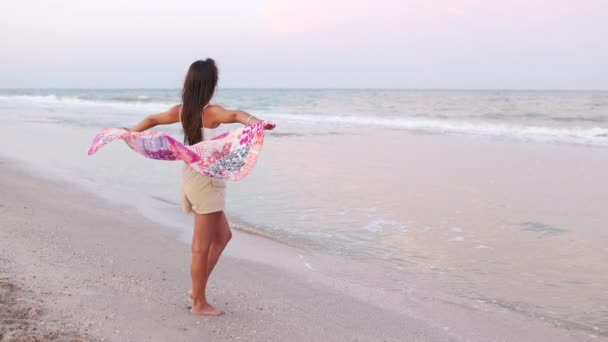 Девушка в летней футболке и шортах идет по пляжу, глядя на горизонт и держа в руках шаль — стоковое видео