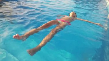 Pembe mayo giymiş bir kız yaz tatilinde temiz su dolu bir havuzda sırt üstü yüzer.