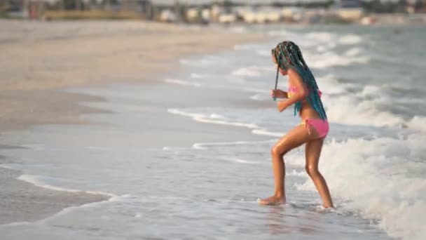 Девушка с африканскими косичками в летнем костюме играет на пляже с раковинами у моря с волнами под солнечным закатом — стоковое видео