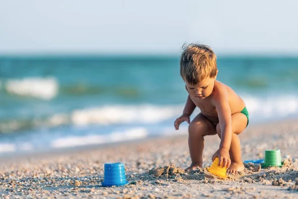 Chico jugando con juguetes en la playa construyendo perlas y torretas sonriendo a alguien detrás de las escenas en las vacaciones de verano —  Fotos de Stock