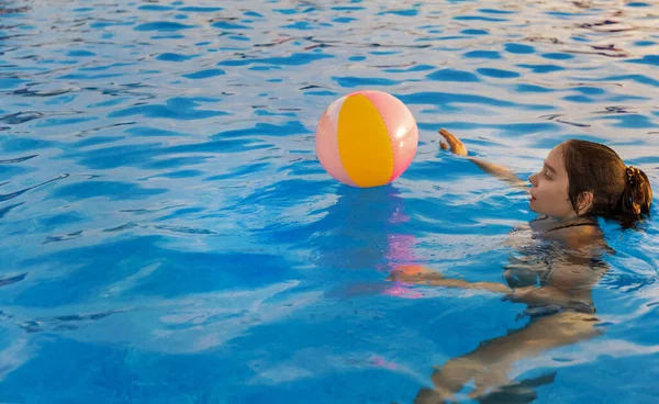 En flicka i en ljus baddräkt simmar med en uppblåsbar boll i en pool med klart vatten en sommarkväll — Stockfoto