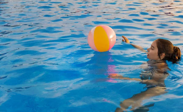 Una chica en un traje de baño brillante nada con una pelota inflable en una piscina con agua clara en una noche de verano — Foto de Stock