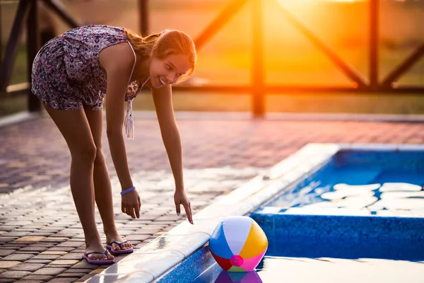 Una chica con el pelo trenzado en un moño en traje brillante juega junto a la piscina con una pelota contra el fondo del sol de verano — Foto de Stock