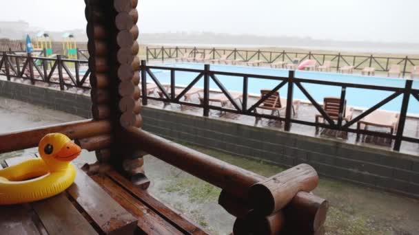 Um pato inflável encontra-se na mesa de madeira no gazebo no centro de recreação contra o pano de fundo da piscina coberta com chuva — Vídeo de Stock