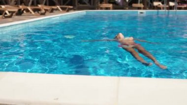 Pembe mayolu ve gözlüklü bir kız havuzda sırtüstü uzanıyor ve yaz tatilinin tadını çıkarıyor.