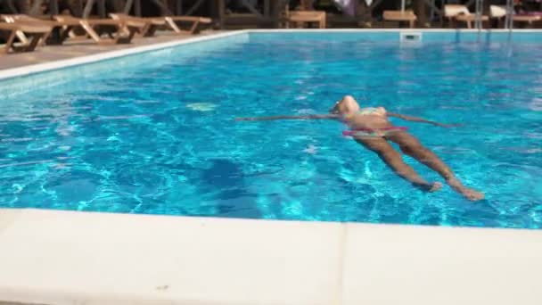 眼鏡をかけたピンクの水着を着た女の子が彼女の背中のプールの水に横たわっており、夏休みを楽しんでいます — ストック動画