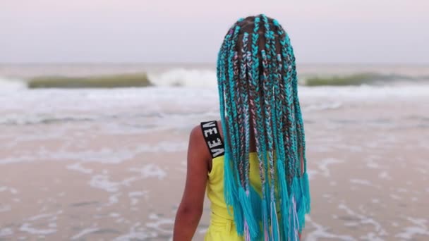 Dziewczyna w letnim garniturze z afrykańskimi warkoczami spogląda na horyzont morza stojąc na plaży — Wideo stockowe
