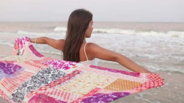 Yaz tişörtlü ve şortlu bir kız sahilde yürüyor ufka bakıyor ve elinde bir şal tutuyor. — Stok video