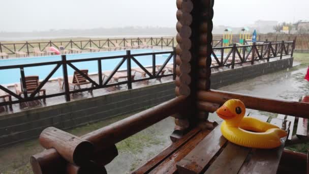 Надувна качка лежить на дерев'яному столі в альтанці в центрі відпочинку на тлі басейну, покритого дощем — стокове відео