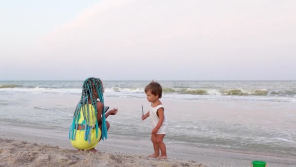 Un niño con una camiseta a rayas baila al son de la música con su hermana trenzas africanas en la playa cerca del mar — Vídeos de Stock