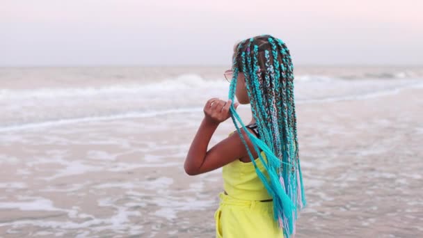 Девушка в летнем костюме с африканскими косичками смотрит на морской горизонт, стоя на пляже — стоковое видео