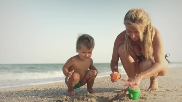 Mamá con gafas juega con un pequeño niño sentado en la playa cerca del mar — Vídeos de Stock