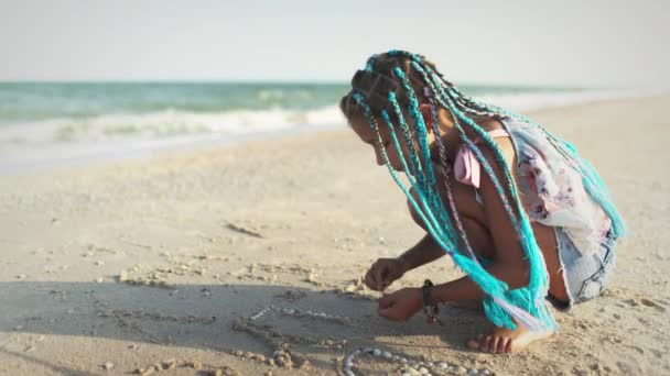 Una chica con trenzas africanas en un disfraz de verano juega en la playa con conchas cerca del mar con olas bajo la puesta de sol — Vídeos de Stock