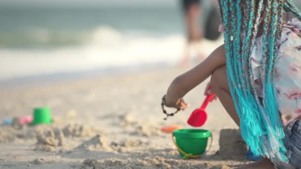 Yazlık kostümü içinde Afrika örgülü bir kız deniz kenarında deniz kabuklarıyla güneşli günbatımında dalgalarla sahilde oynuyor. — Stok video