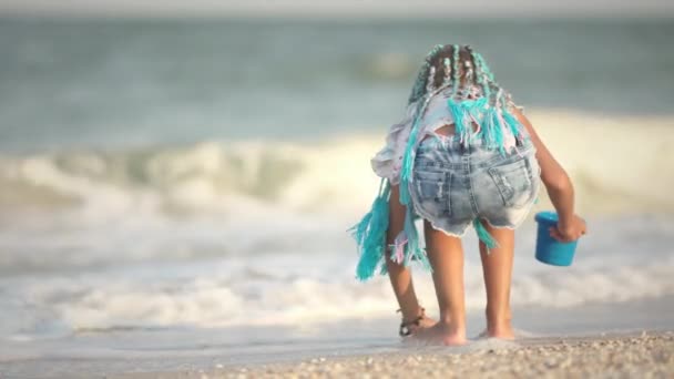 Девушка с африканскими косичками в летнем костюме играет на пляже с раковинами у моря с волнами под солнечным закатом — стоковое видео