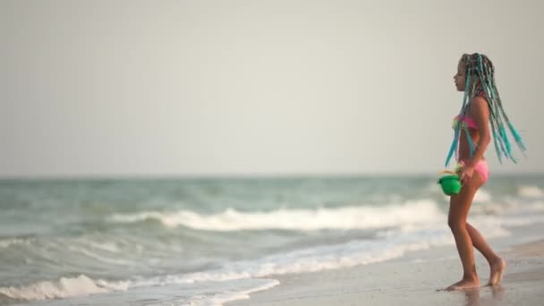Dziewczyna z afrykańskimi warkoczami w letnim kostiumie bawi się na plaży z muszlami w pobliżu morza z falami pod słońcem — Wideo stockowe