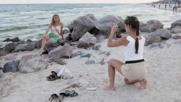 A girl with a scythe and a mobile phone takes pictures of her mother who is sitting on a stone on the sea coast — Vídeos de Stock