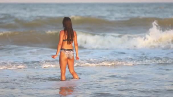 Egy laza hajú lány hullámokkal sétál a tengeren, és élvezi a napot. — Stock videók