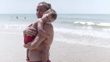 Yaz bikinili anne, deniz kenarında tatilde uyumak isteyen bir bebeği kucağında tutuyor.