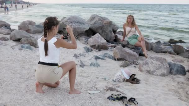 Девушка с косой и мобильным телефоном фотографирует свою мать, сидящую на камне на берегу моря — стоковое видео