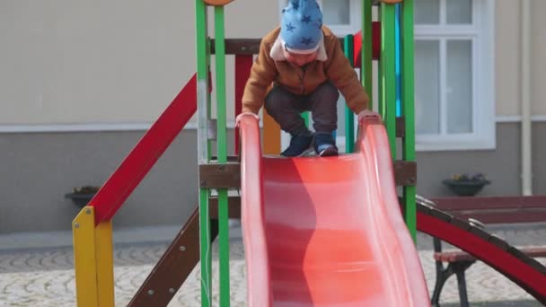 The boy plays on the playground and rides a slide in autumn weather — Videoclip de stoc