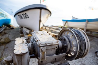 Geniş bir taş ve beton iskele üzerinde gri bulutlu bir gökyüzüne karşı çeşitli küçük demirli tekne ve teknelerle güvenilir, ince, siyah bir iple eski paslı bir vinç.