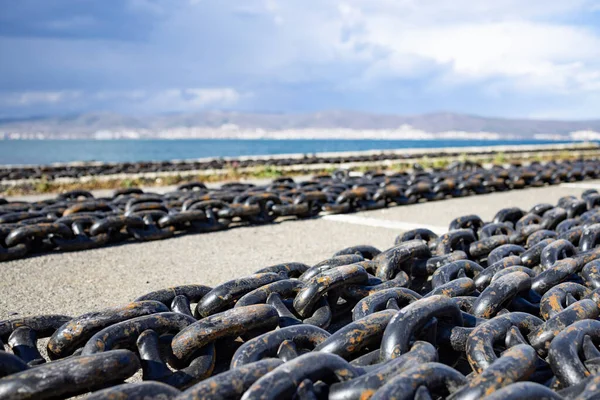 Large long sea chains made of rusty old dark gray metal for ships and boats lie on a wide stone empty pier, against the background of a cloudy blue spacious sky and a quiet calm blue Black Sea