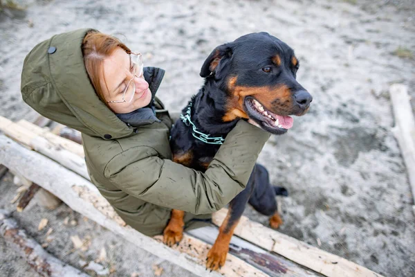 在狂野的沙滩上 寒冷多云的天气里 穿着温暖衣服 和蔼而快乐的女主人姑娘鬼混在一起 与她可笑的大忠实的狗朋友罗特韦勒品种拥抱在一起 — 图库照片