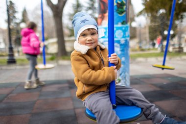Mavi şapkalı ve sıcak elbiseli neşeli aktif bir çocuk boş bir sonbahar parkındaki çocuk parkında boş zamanlarını parlak bir çocuk parkında geçirir ve bulutlu havada mavi, modern bir salıncakta gezer.