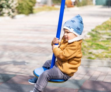 Mavi şapkalı ve sıcak elbiseli neşeli aktif bir çocuk boş bir sonbahar parkındaki çocuk parkında boş zamanlarını parlak bir çocuk parkında geçirir ve bulutlu havada mavi, modern bir salıncakta gezer.
