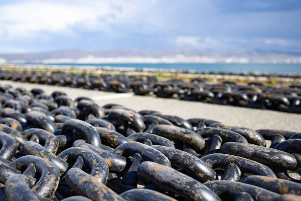 Large long sea chains made of rusty old dark gray metal for ships and boats lie on a wide stone empty pier, against the background of a cloudy blue spacious sky and a quiet calm blue Black Sea