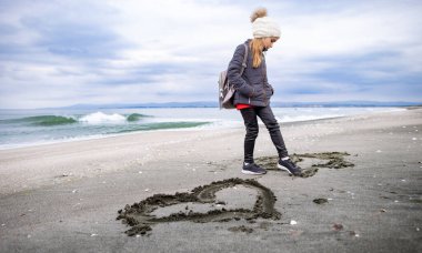 Koyu renk elbiseli, hayalperest bir kız ve büyük bir sırt çantalı beyaz bir şapka boş kumlu plajımda yürüyor ve ıslak kumlara çocuk çizimleri ve sembolleri çiziyor.