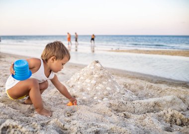 Neşeli, hevesli bir çocuk kumlu bir sahilde oyuncaklarıyla oynuyor. Boncuk ve taret yapıyor. Parlak güneşli bir yaz tatilinde sahne arkasında birine gülümsüyor.