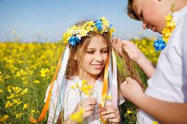Önemseyen ağabeyi, parlak Ukrayna çiçek çelenkleriyle çok renkli ince kurdeleler örüyor. Küçük, gülümseyen kız kardeşin başında. Açık mavi gökyüzünün altında bitmeyen kolza tohumu tarlasında.