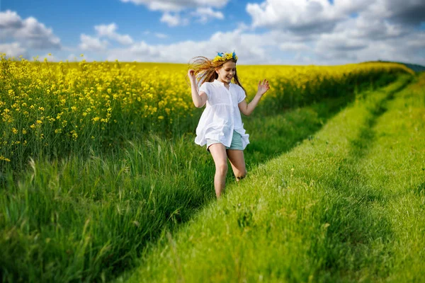 白いドレスと明るいウクライナの色の花輪で素敵な精力的な夢の十代の少女は 青い曇った空に対して 黄色の開花菜の花畑と緑の春の無限の牧草地を通って実行されます — ストック写真