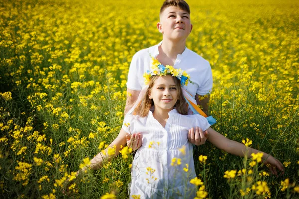 Big strong older teenage brother catches his little cheerful happy sister and bright Ukrainian flower wreath that falls into his hands, in rapeseed yellow flowering field under blue clear sky