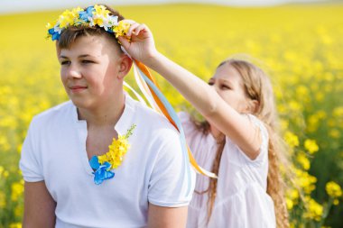 Beyaz elbiseli neşeli kız kardeş Ukrayna 'da çok renkli yaz kurdeleleri takıyor. Genç kardeşinin başında sarı tarlalar ve açık gökyüzü var.