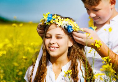 Neşeli gülen genç adam parlak Ukrayna çiçek çelenkleriyle açık gökyüzüne ve çiçek açan sarı tarlalara gülümseyen şirin kız kardeşine çok renkli uzun kurdeleler fırlatıyor.