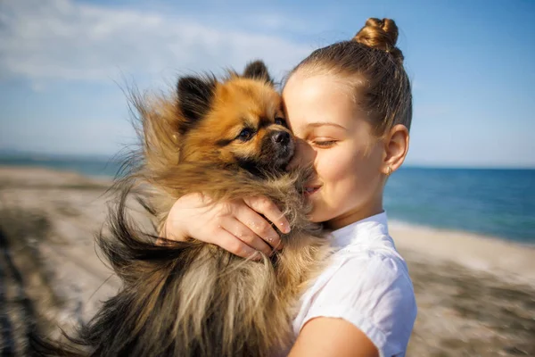 Saçları düzgün örülmüş sarı saçlı, beyaz tişörtlü, neşeli gülüşlü, pomeranian köpeğini öpen, altın sarısı uzun saçlı, Karadeniz kıyısında, güneşli rüzgarlı havada.