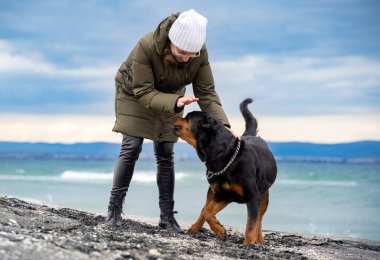 Sıcak giysili mutlu yetişkin ev sahibesi kadın, kumlu bir sahilde soğuk bulutlu bir havada Rottweiler cinsinin sadık ve komik köpeğiyle oynaşıyor.