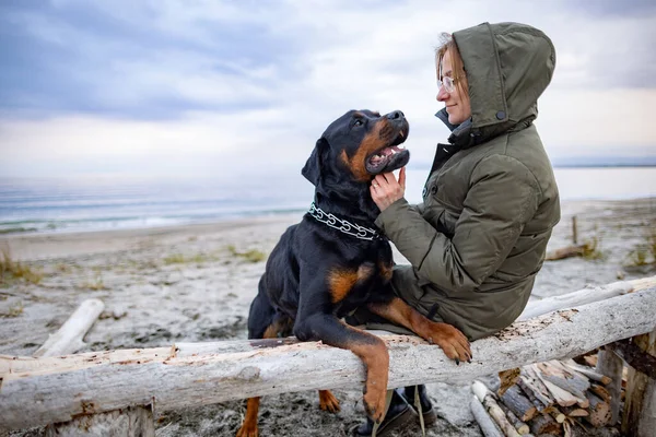 在狂野的沙滩上 寒冷多云的天气里 穿着温暖衣服 和蔼而快乐的女主人姑娘鬼混在一起 与她可笑的大忠实的狗朋友罗特韦勒品种拥抱在一起 — 图库照片