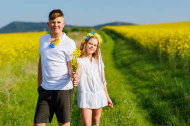 Mutlu kaygısız yerli çocukların ön görüntüsü: Genç erkek kardeş ve küçük kız kardeş, bulutlu gökyüzüne karşı sarı açan kolza tohumu tarlalarıyla çevrili yeşil çimlerle uzaklarda yürüyorlar.