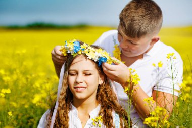Neşeli gülen genç adam parlak Ukrayna çiçek çelenkleriyle açık gökyüzüne ve çiçek açan sarı tarlalara gülümseyen şirin kız kardeşine çok renkli uzun kurdeleler fırlatıyor.